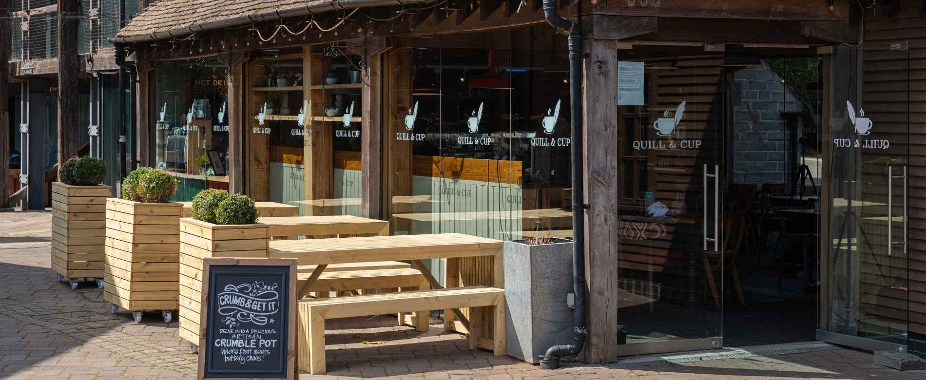 An exterior shot of The Quill and Cup Café in Stratford-upon-Avon. There is a wooden seating area next to the large glass windows and a sign to display their Crumble Pots